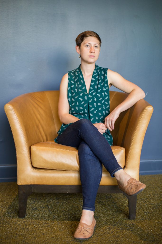 Full-length indoor portrait of a professional writer wearing a green shirt, seated in an oversized taupe leather chair against a blue backdrop.