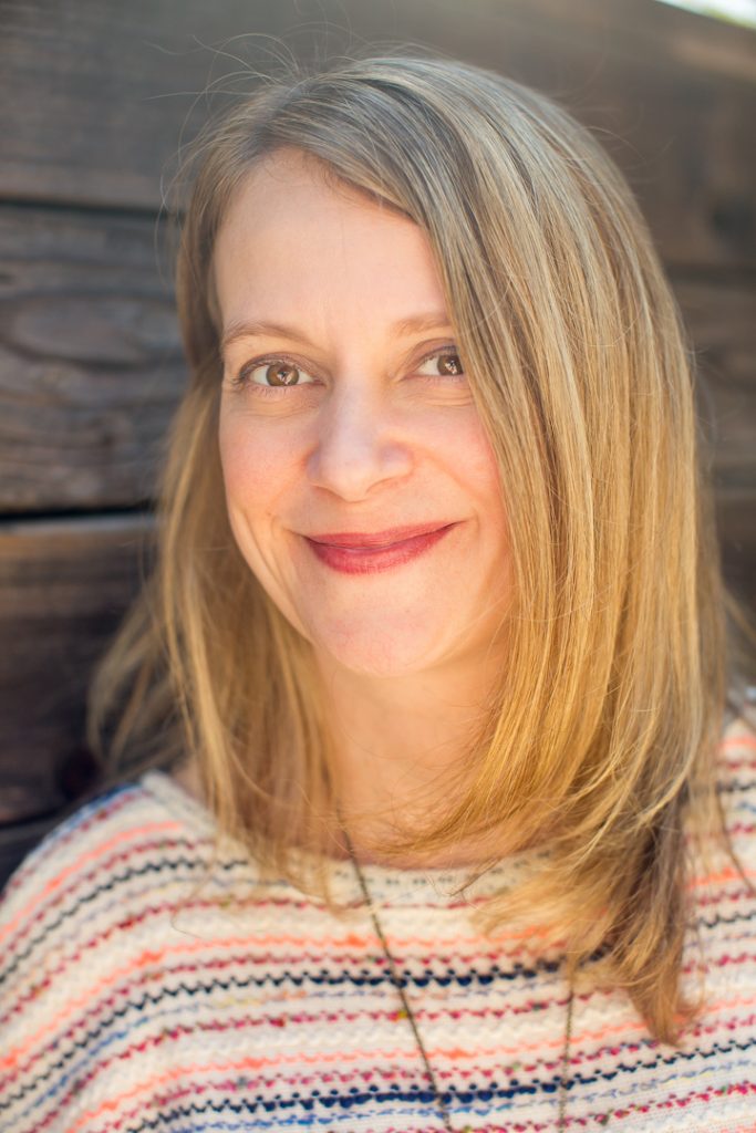 A warmly lit headshot of a professional artist smiling against a wooden backdrop.