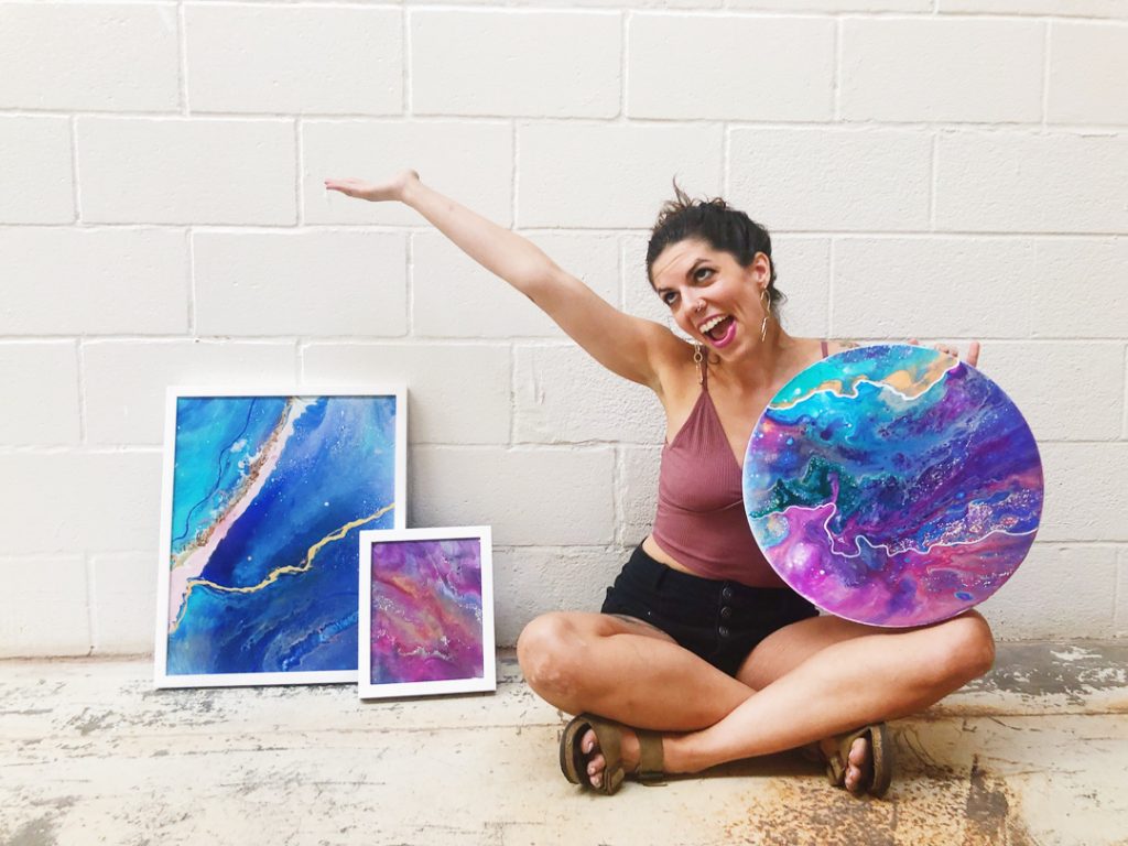 Courtney Potter sitting on the floor cross-legged next to 2 framed paintings, holding a round canvas fluid painting from her Voyage Collection.