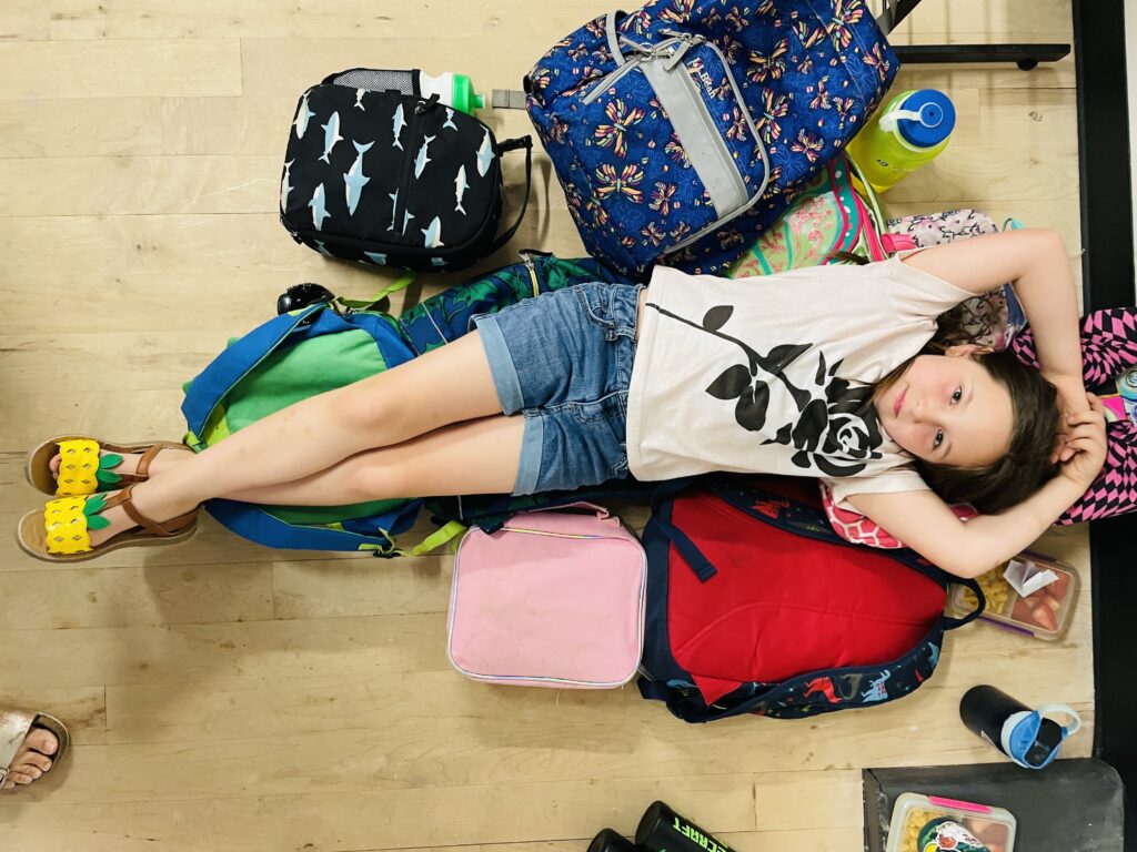 A young girl lays on a pile of colorful backpacks during art camp.