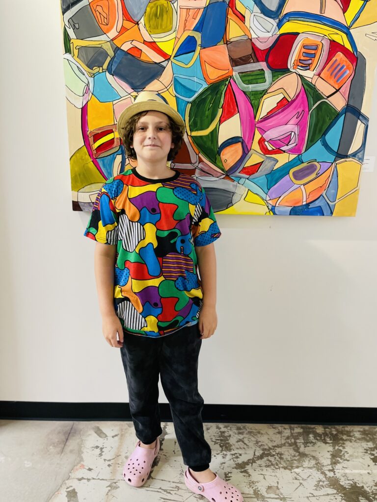 A middle school-aged boy wearing a hat and a colorful abstract shirt poses in front of a giant abstract painting that matches his shirt at art camp