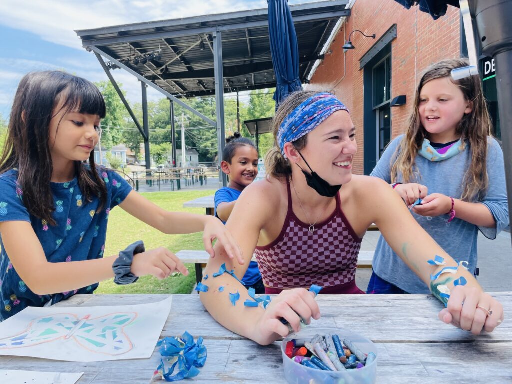 Three kids playfully put tape on a teaching artist during art camp.