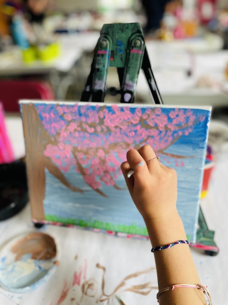 A camper wearing two handmade friendship bracelets paints cherry blossoms on her canvas during a spring mini art camp