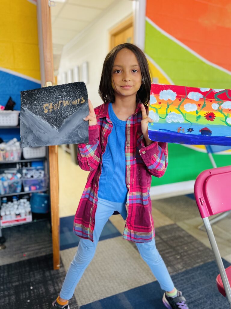 A girl wearing red flannel shows off her artwork from camp against a rainbow wall